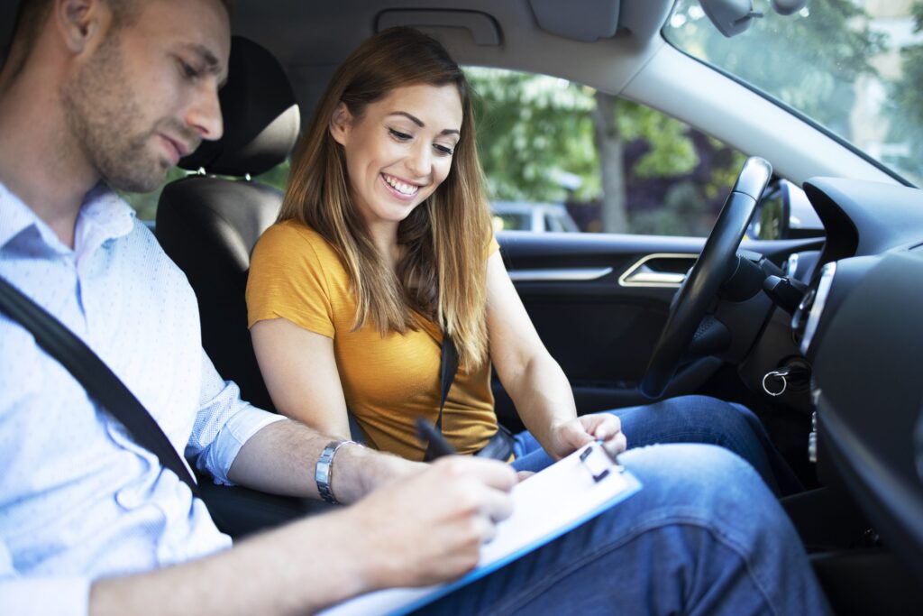 Casal sorridente dentro de um carro, a mulher está segurando uma prancheta enquanto o homem escreve. Ambos parecem estar se divertindo durante uma atividade de aprendizado ou instrução.
